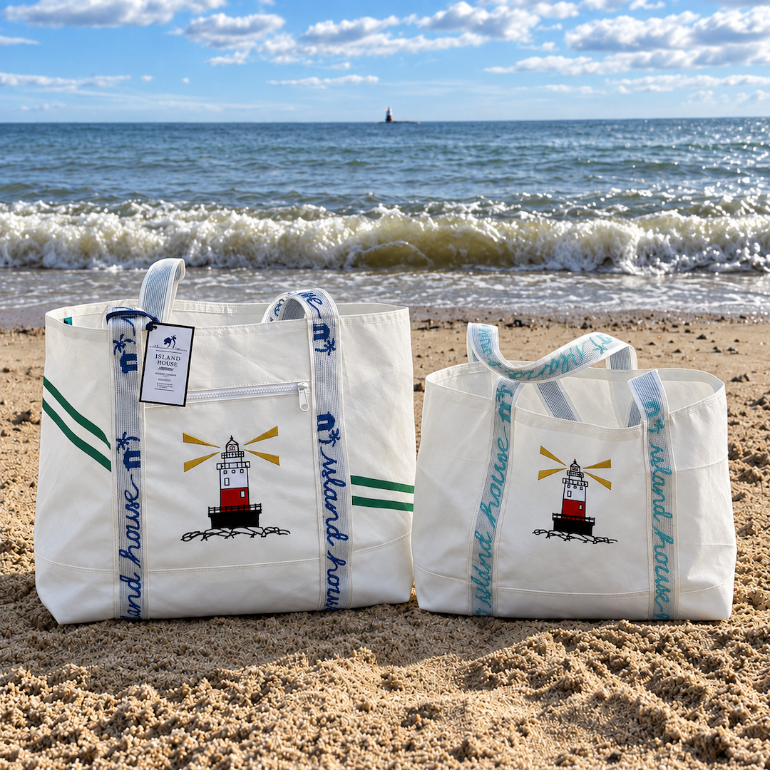 The front of a two totes made of white reclaimed sailcloth. A lighthouse is embroidered on the middle of the front of each bag. The left bag is a large size and the right bag is a medium. The bags are sitting side-by-side on the beach at the Wee Burn Beach Club.
