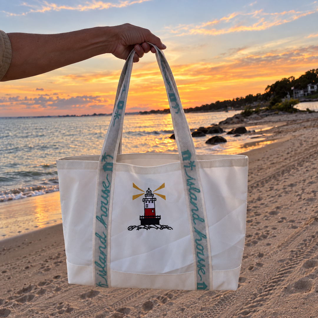 The front of a medium tote made of white reclaimed sailcloth. The straps are made of sail tie webbing and are white with aqua blue lettering that reads "Island House". A lighthouse is embroidered on the middle of the front. A hand is holding the bag on the shore of the Wee Burn Beach Club.